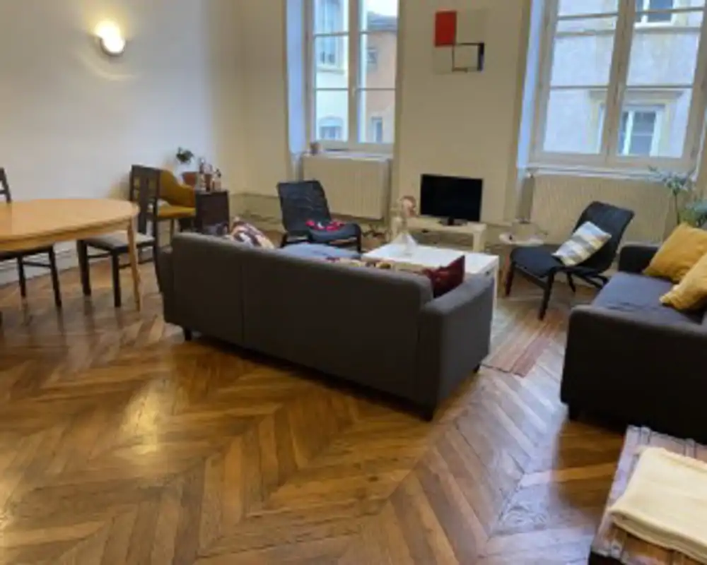 Spacious, well-lit living room with a central sofa arrangement, armchairs, TV and dining area; large windows and warm parquet flooring create a welcoming atmosphere.