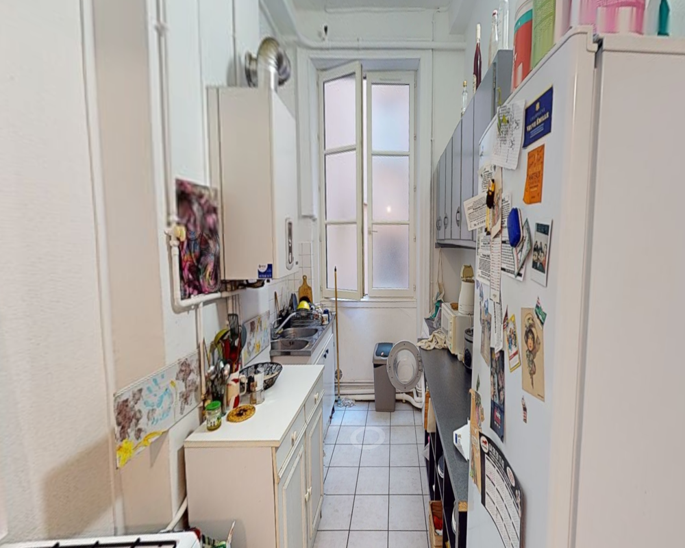 Cuisine en couloir étroite avec une fenêtre au fond, plan de travail et évier à gauche et un réfrigérateur et des rangements à droite ; des éléments personnels comme des magnets, petits appareils et ustensiles sont visibles.