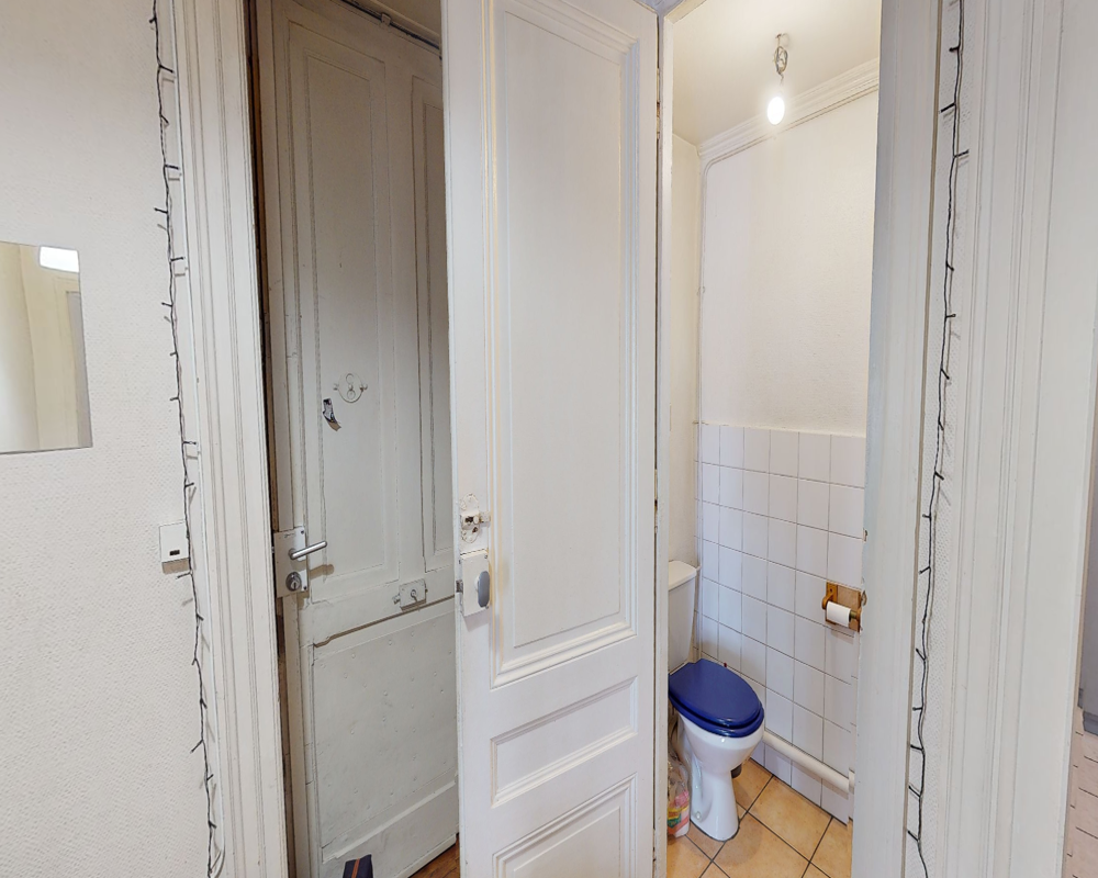 Petite salle de bains avec WC à abattant bleu, demi-mur carrelé blanc et sol carrelé ; porte ouverte sur un espace étroit avec éclairage simple.