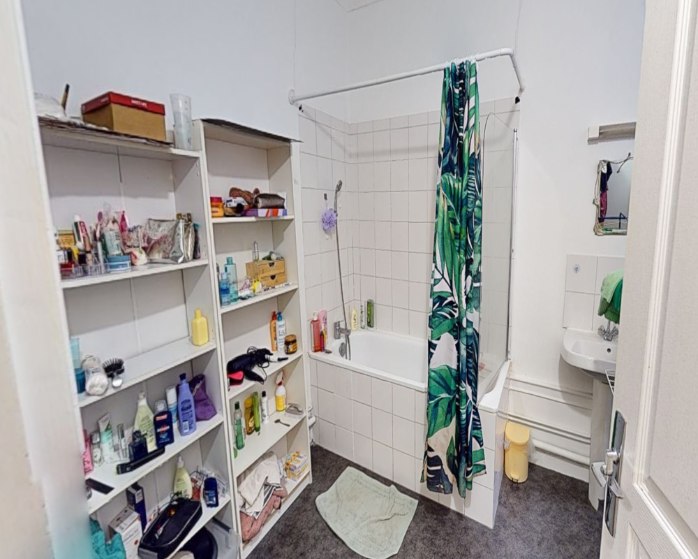Salle de bain compacte avec baignoire carrelée et rideau de douche, douche murale, lavabo et étagères ouvertes pour les produits — espace lumineux et fonctionnel adapté aux annonces locatives.