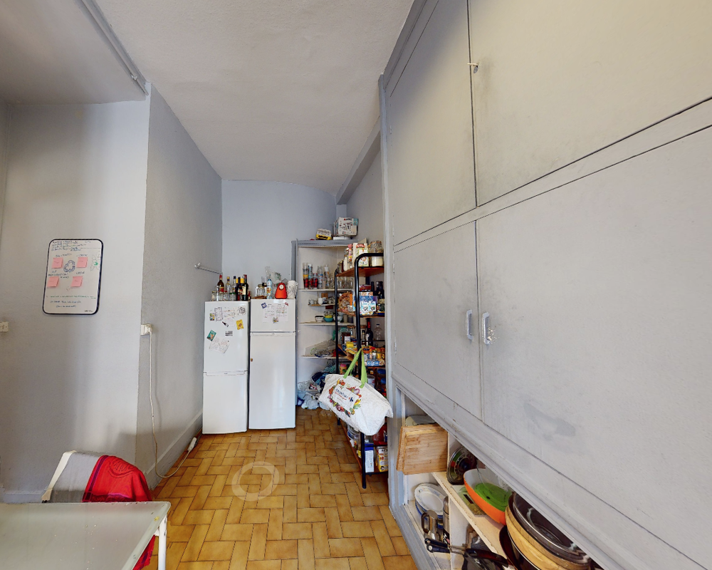 Área de cocina/despensa funcional con dos frigoríficos, estanterías abiertas con alimentos y utensilios, y armarios empotrados en la pared derecha. Espacio práctico y vivido, apto para el uso diario.