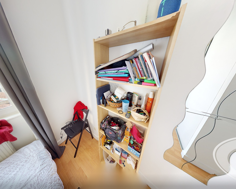 Small bedroom corner with a visible bed at the left, a tall wooden shelving unit filled with personal items, and a window providing bright natural light. The space feels lived-in and functional with moderate clutter.