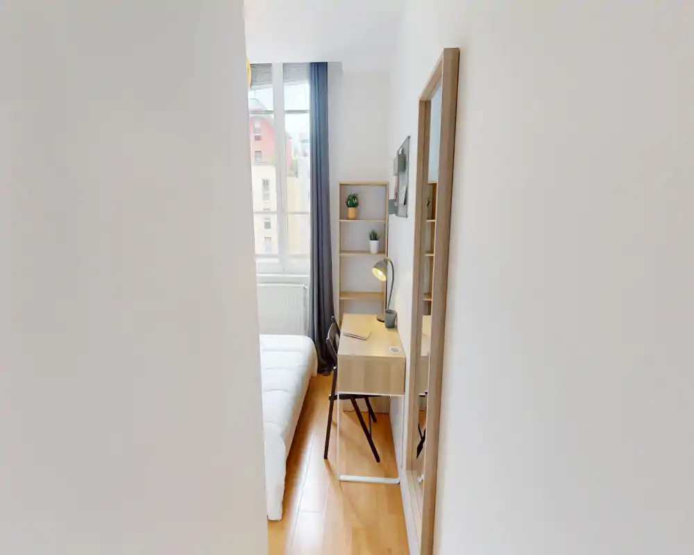 Bright, compact bedroom with a neatly made bed, a small wooden desk with lamp and chair, and a tall window providing natural light. The neutral palette and minimalist shelving create a calm, functional space ideal for short stays.