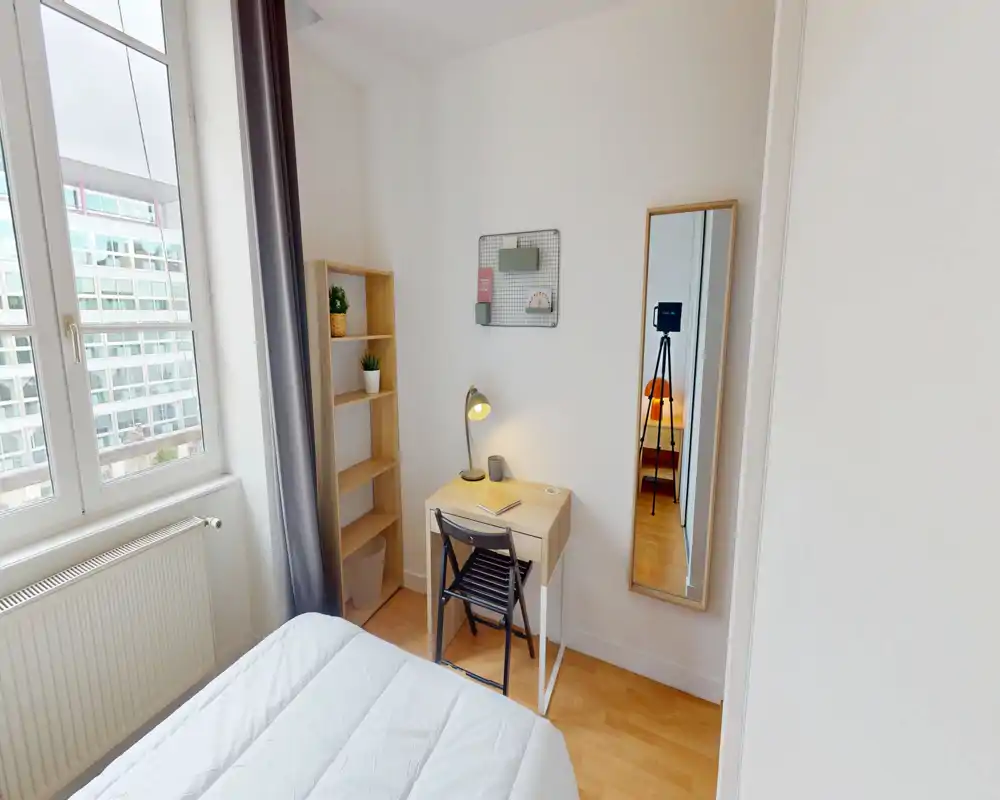 Bright, neatly staged small bedroom with a white bed, a compact desk and shelving by a large window letting in abundant natural light.