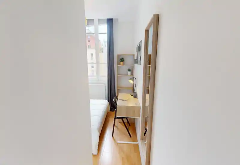 Bright, compact bedroom with a neatly made bed, a small wooden desk with lamp and chair, and a tall window providing natural light. The neutral palette and minimalist shelving create a calm, functional space ideal for short stays.