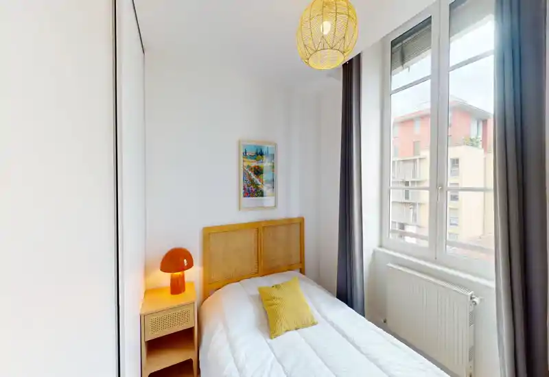 Bright, modern single bedroom with a rattan headboard, cozy yellow accent pillow and a bedside lamp. Large window provides ample natural light and neutral walls make the room feel airy.