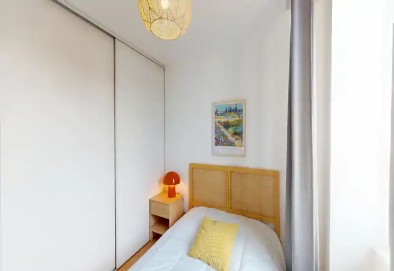 Compact, bright bedroom with a woven headboard, bedside table and warm bedside lamp. Natural light from a large window and soft textiles create a welcoming atmosphere.