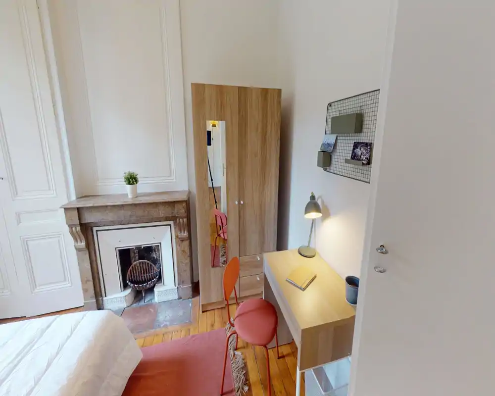 Camera da letto luminosa e accogliente con letto ben visibile, armadio in legno con specchio, scrivania e sedia accanto a un camino decorativo — ideale per un soggiorno confortevole.