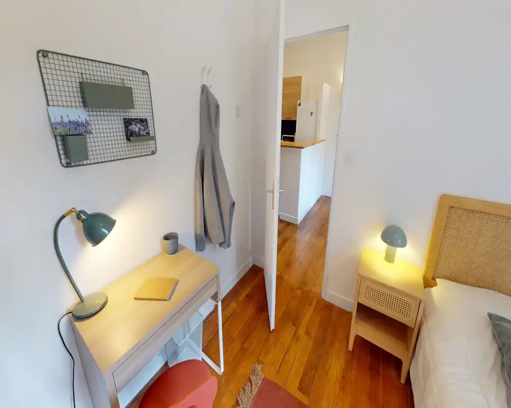 Angolo di camera luminoso e accogliente con testiera in legno, comodino e una piccola scrivania con lampada. Pavimento in legno caldo e elementi decorativi semplici rendono lo spazio ideale per una persona o come camera ospiti.