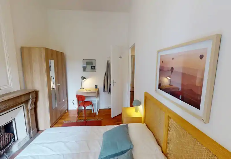 Camera da letto ben arredata con comodo letto matrimoniale in primo piano, comodino con lampada, scrivania con sedia e armadio — ambiente luminoso e caldo con pavimenti in legno.