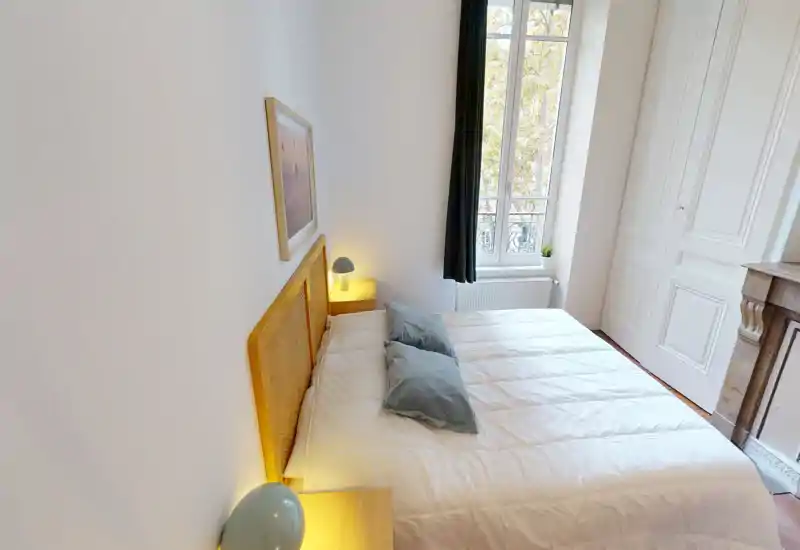 Camera da letto luminosa e di stile con grande letto matrimoniale, cuscini morbidi e lampade da comodino calde. La finestra alta garantisce luce naturale e un’atmosfera rilassante.