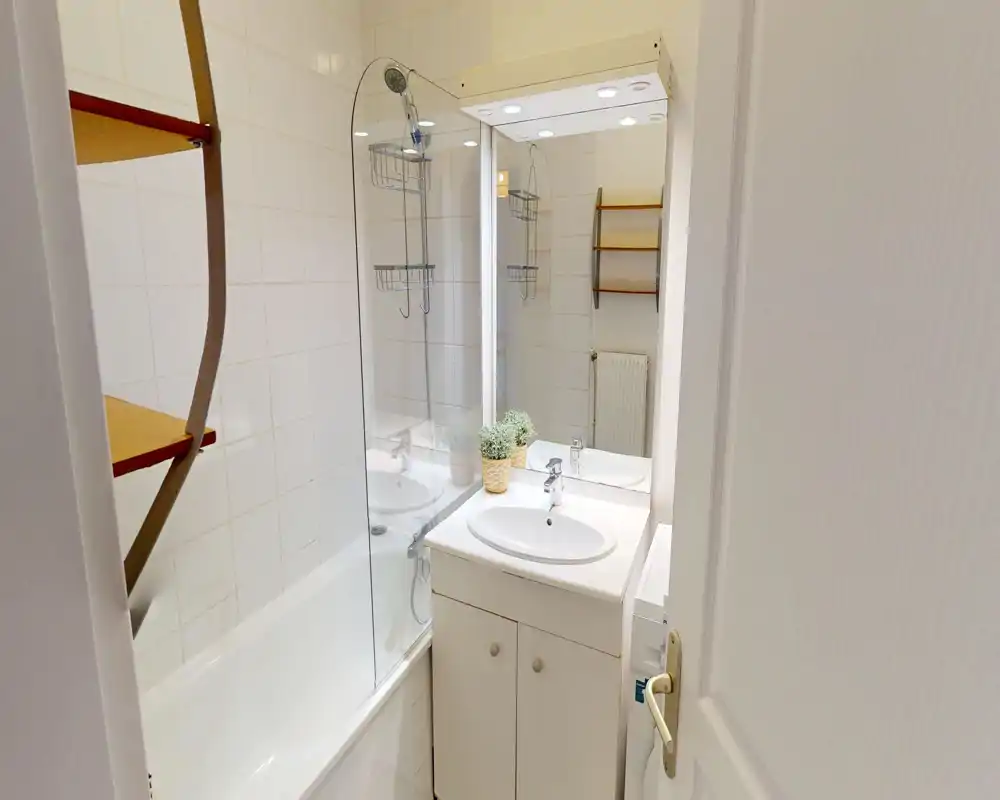 Bagno compatto e ben illuminato con vasca, parete doccia in vetro, lavabo con mobile sottostante e specchio pensile; piastrelle bianche per un aspetto pulito e luminoso.