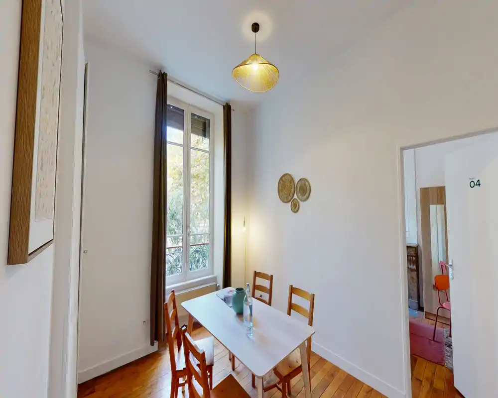 Soggiorno-pranzo luminoso e semplice con un tavolo in legno e quattro sedie davanti a una grande finestra. Luce naturale e pavimento in legno creano un'atmosfera accogliente per i pasti.