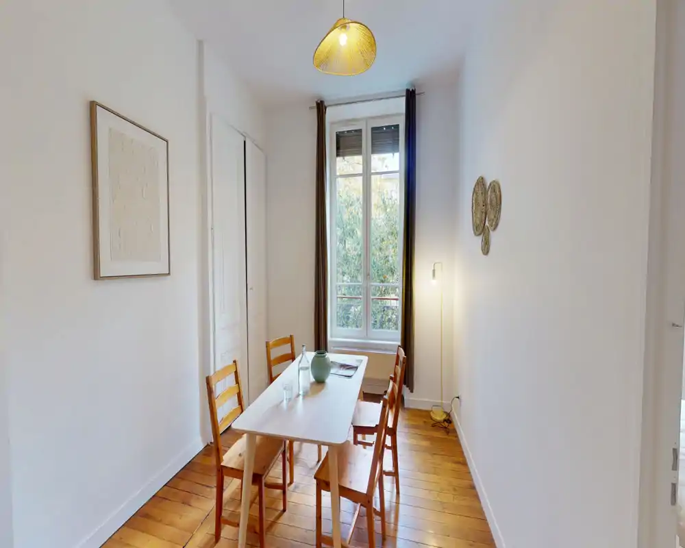 Sala da pranzo compatta e luminosa con un semplice tavolo in legno e quattro sedie, luce naturale da una finestra alta e pavimento in legno che rende l'ambiente accogliente.