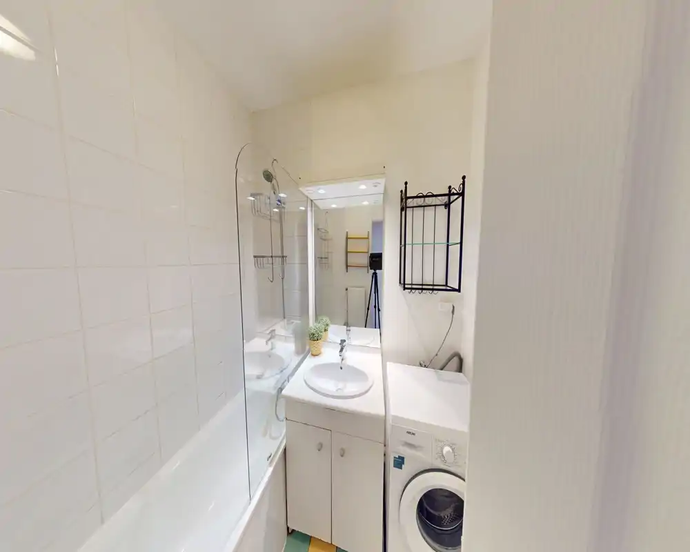 Bagno compatto e ben illuminato con vasca e paratia in vetro, lavabo con mobiletto e lavatrice a carica frontale. Piastrelle neutre e sanitari bianchi creano un ambiente luminoso e funzionale.