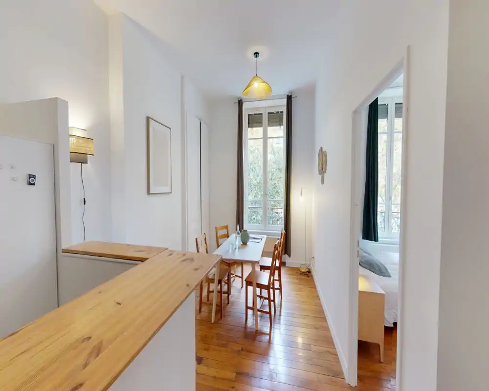Zona pranzo luminosa e semplice con tavolo e sedie in legno vicino a una finestra alta, pavimenti in legno caldo e arredi minimal che creano un'atmosfera accogliente.