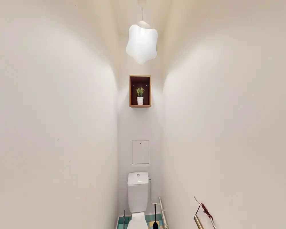 WC compatto e ben illuminato con wc moderno, mensola minimal e lampada a sospensione decorativa; pareti bianche luminose per un senso di pulizia.