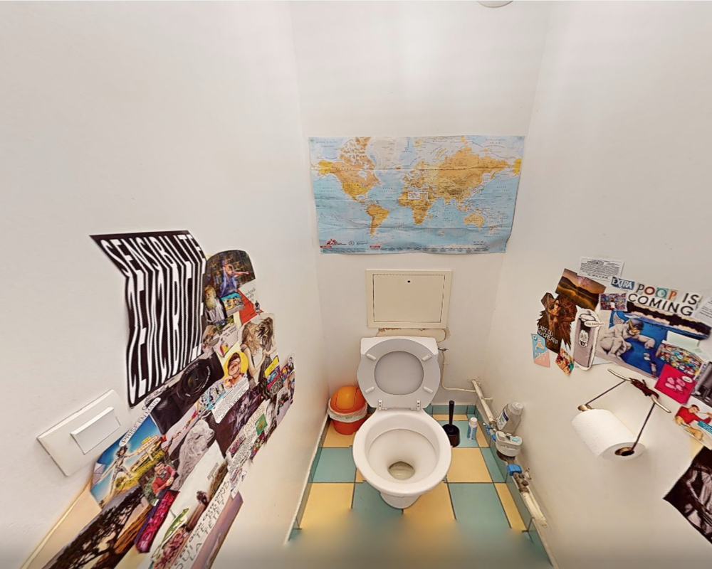 Piccolo bagno privato con wc a parete, pavimento piastrellato colorato e poster decorativi alle pareti. Layout compatto con scopino e rotolo di carta igienica a portata di mano.