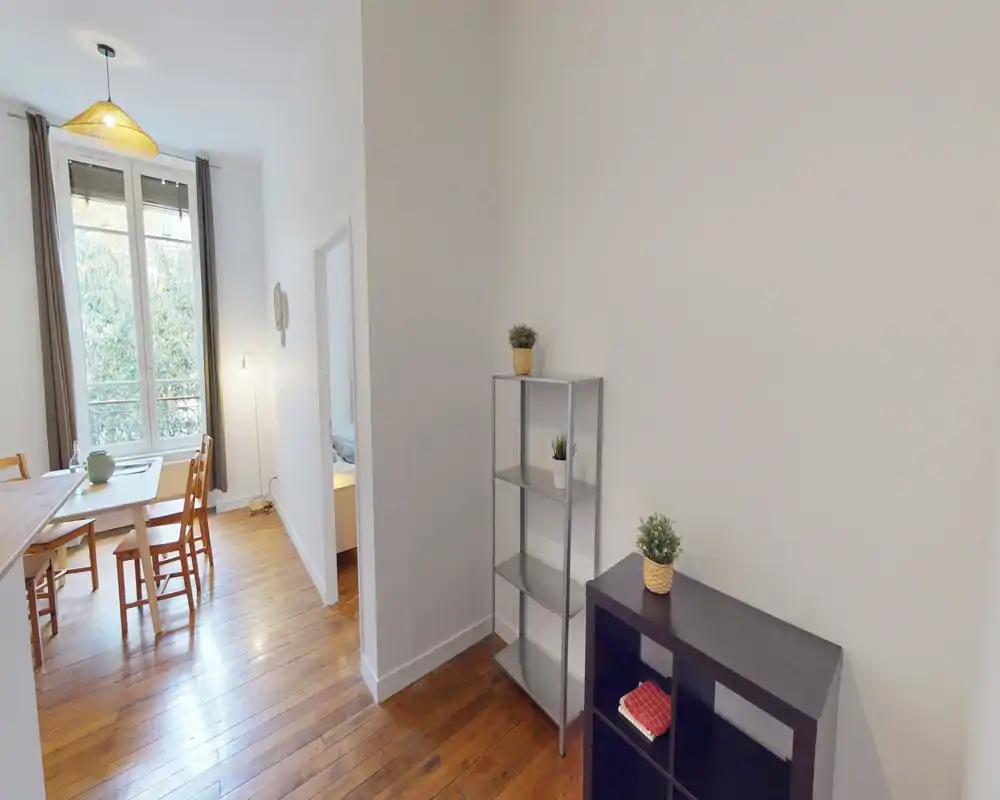 Zona giorno luminosa e semplice con pavimenti in legno, tavolo da pranzo vicino a una grande finestra e scaffalature minimaliste — perfetta per un appartamento in città.