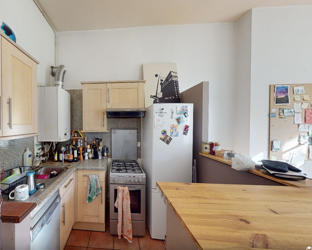 Cucina compatta e vissuta con mobili in legno, piano cottura a gas indipendente, frigorifero con calamite e bancone in legno — luce naturale a destra e disposizione pratica ideale per un piccolo appartamento.