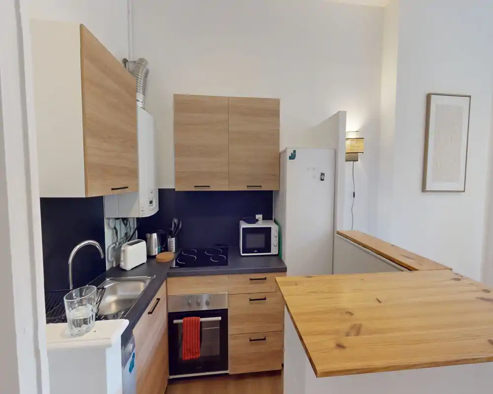 Cucina compatta e moderna con mobili effetto legno, piano a induzione, forno integrato e piccolo bancone — luminosa e ordinata per un uso efficiente.
