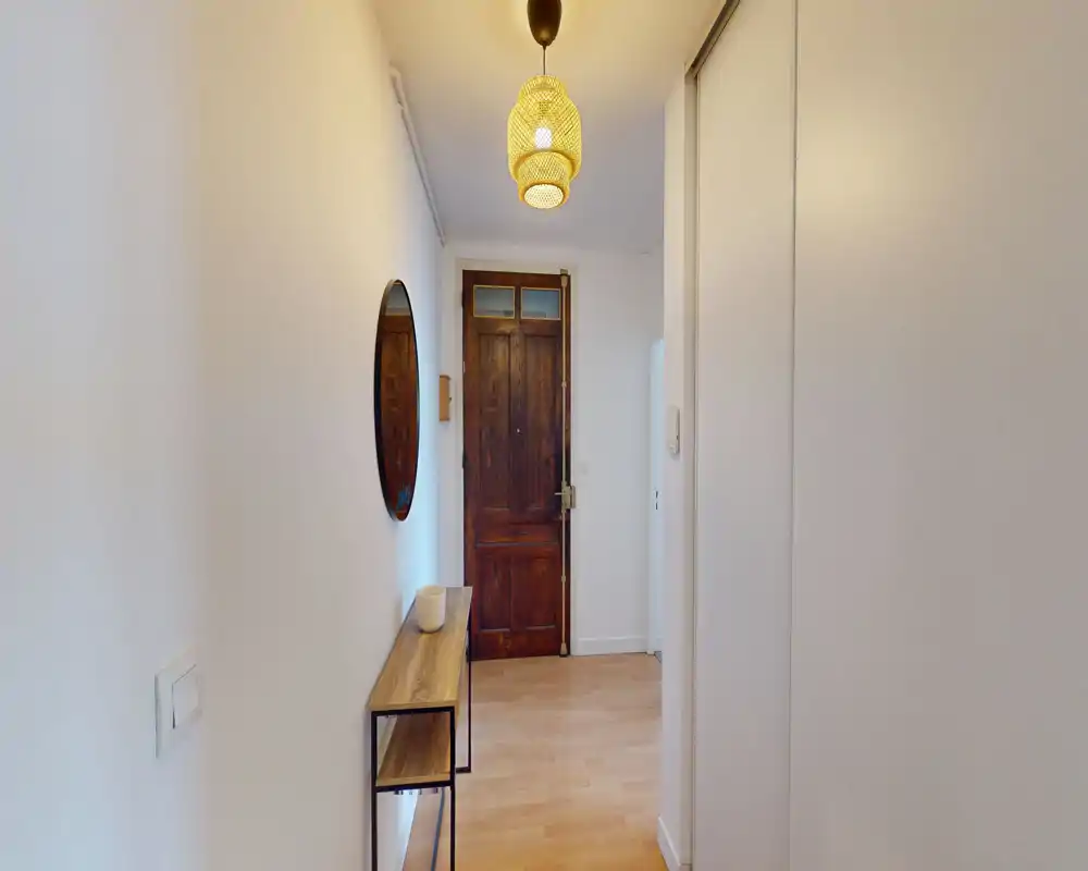 Ingresso luminoso e stretto con porta d'ingresso in legno, un sottile tavolino consolle e uno specchio rotondo; lampada a sospensione calda e pavimento in legno chiaro creano una prima impressione accogliente.