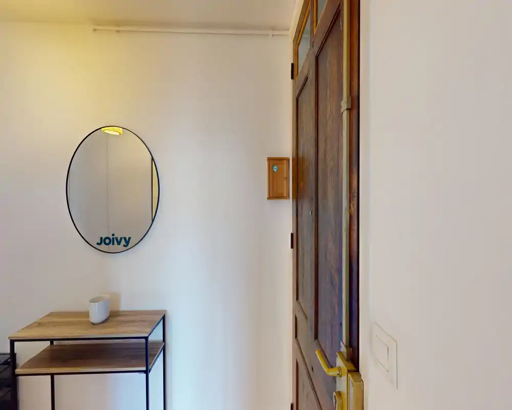 Ingresso piccolo e luminoso con porta in legno, specchio tondo a parete e un sottile tavolino consolle — ordinato e ben illuminato.