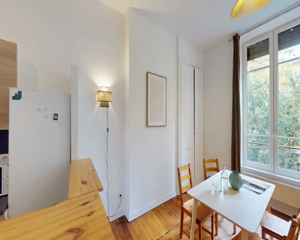 Piccola zona pranzo luminosa vicino a una grande finestra con tavolo e sedie in legno semplici, vaso decorativo e luce naturale — ideale per pasti accoglienti.