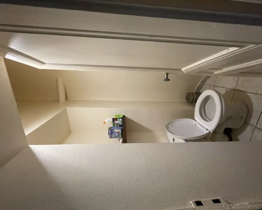 WC compact avec toilette standard, petite étagère au-dessus avec produits d'entretien, sol carrelé et murs neutres — adapté pour une personne.