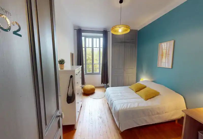 Bright, well-styled bedroom with a double bed against a teal accent wall, natural light from a tall window, wooden floors and cozy accents.