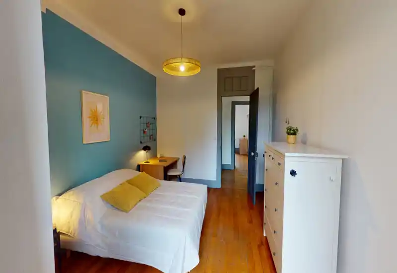Bright, well-staged bedroom with a white bed against a teal accent wall, wooden flooring and a small desk at the foot of the bed — cozy and inviting for guests.