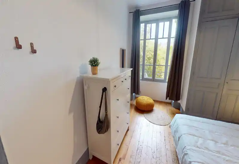 Bright furnished bedroom with wooden floors, a comfortable bed, chest of drawers and large window providing generous natural light — cozy, minimalist decor ideal for restful stays.