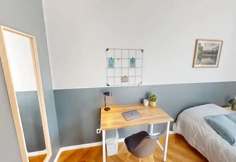 Bright, modern bedroom corner with a neatly made bed, compact wooden desk and chair — ideal for a single or student rental. Neutral tones, parquet flooring and simple decor create a calm, functional space.