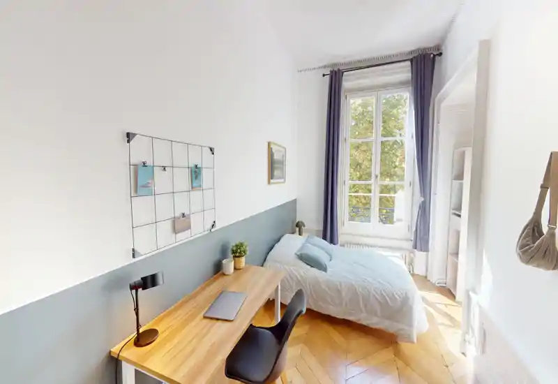 Bright, cosy single bedroom with a comfortable bed next to a large window, a wooden desk for work, and tasteful minimal decor. Natural light and parquet flooring give the room a warm, airy feel.