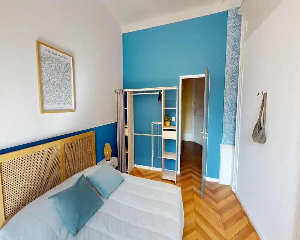 Bright, stylish bedroom with a rattan headboard, blue accent wall and a neatly made bed; hardwood herringbone flooring and an open wardrobe create a fresh, welcoming space.