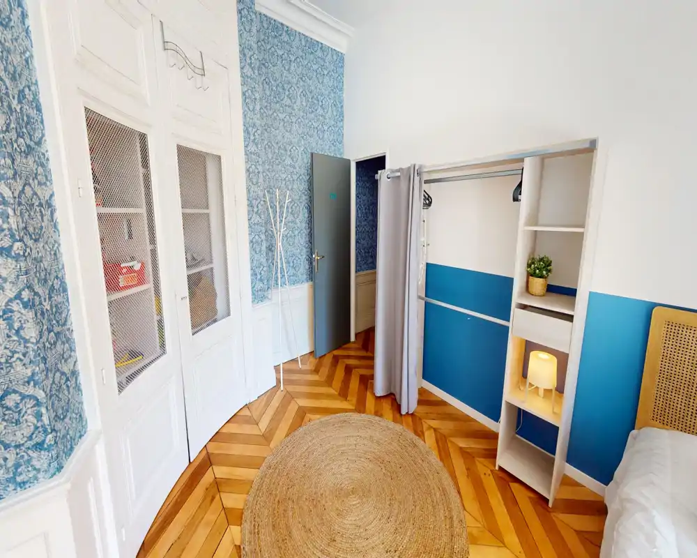 Bright, stylish bedroom corner with a rattan headboard, open wardrobe shelving, decorative blue wall panels and chevron wood floors; cozy round jute rug and soft bedside lamp create a welcoming atmosphere.