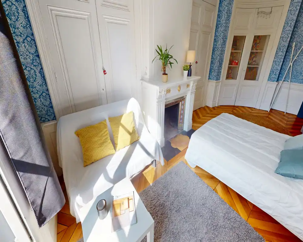 Sunny, elegant bedroom with a comfortable double bed, sofa with cushions, decorative fireplace and wooden herringbone floors — bright and welcoming space ideal for relaxing.