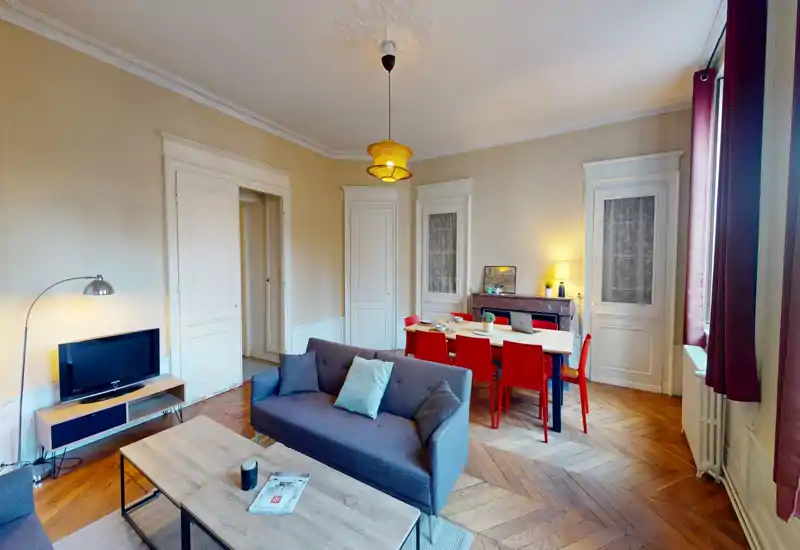 Séjour lumineux et soigné avec canapé gris, table basse, coin TV et espace repas aux chaises rouges — éclairage chaleureux et parquet en chevrons.