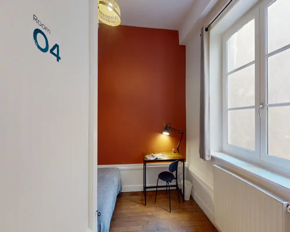 Cozy single bedroom with a warm terracotta accent wall, a small desk with lamp by the window and a partially visible bed — bright natural light and wooden flooring.