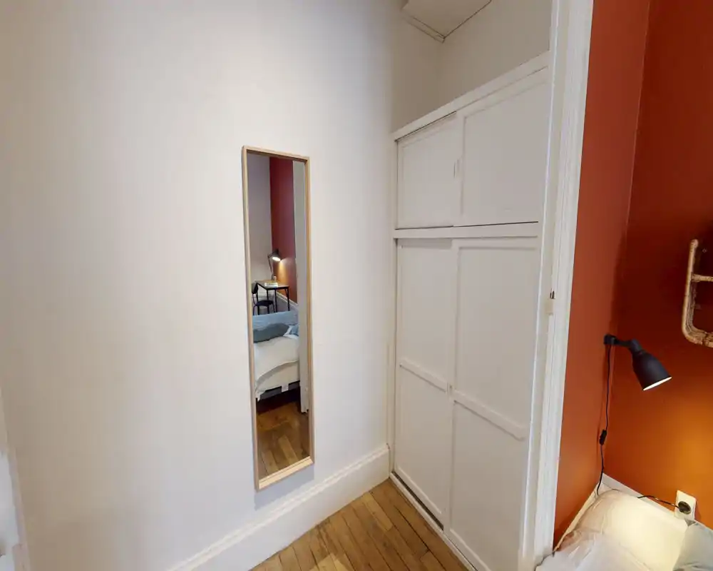 Compact bedroom corner with warm terracotta accent wall, bedside lamp, and partial bed visible; mirror reflects additional bed area and a small bedside table, creating a cozy, well-lit nook.