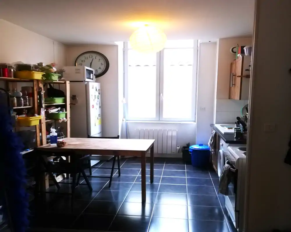Functional kitchen with a wooden dining table, shelving with storage bins, fridge and sink area; large window provides strong natural light and the space feels lived-in.
