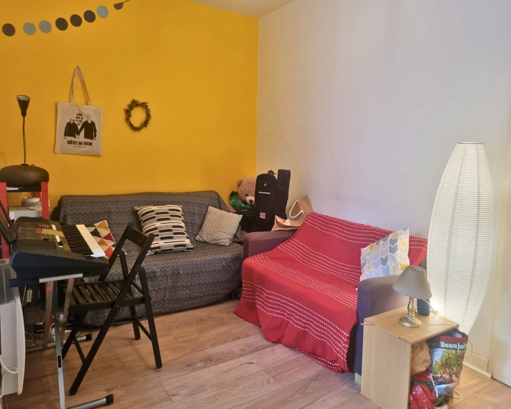 Cozy living area with two sofas, cushions and a keyboard workstation against a bright yellow accent wall. Soft lighting and layered textiles create a warm, lived-in atmosphere.