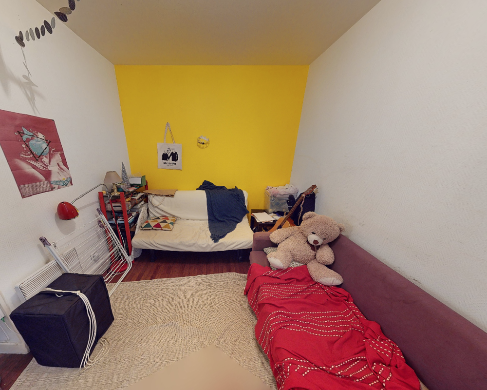 Cozy living area with two sofas, colorful textiles and a yellow feature wall. The space includes decorative cushions, a rug and personal items that create a warm, lived-in atmosphere.