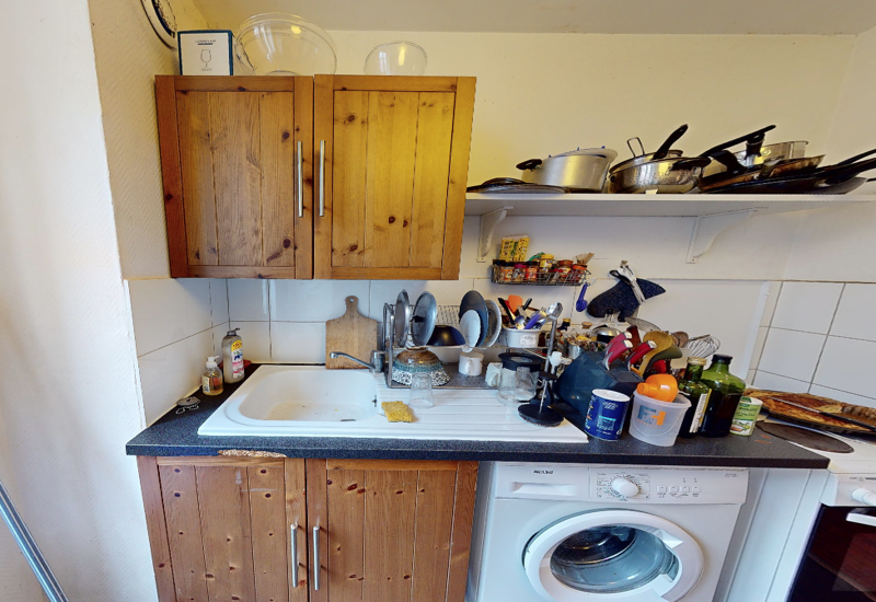 Cucina compatta con pensili in legno, lavello e piano di lavoro con piccoli elettrodomestici; lavatrice visibile e mensole aperte con pentole. Ambiente funzionale e ben illuminato con dettagli casalinghi.