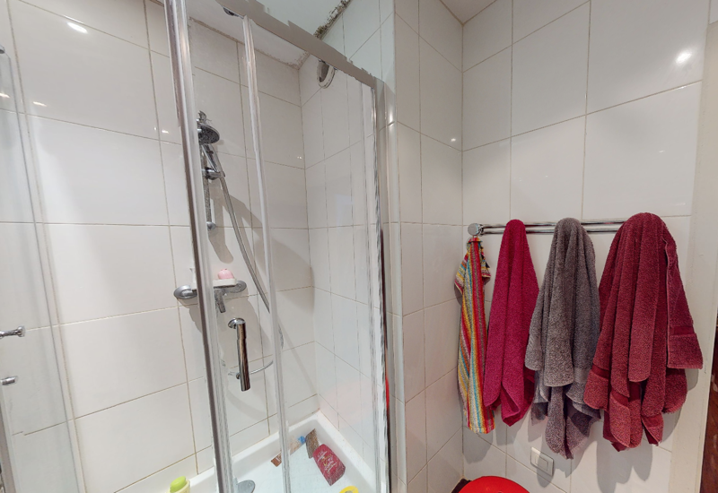 Bagno piastrellato compatto con box doccia in vetro, rubinetteria cromata e porta asciugamani con asciugamani colorati.