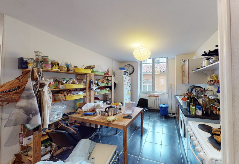 Cucina ben illuminata e funzionale con scaffalature a vista, un tavolo centrale in legno e elettrodomestici sulla parete destra — uno spazio vissuto, ideale per studenti o vita in città.
