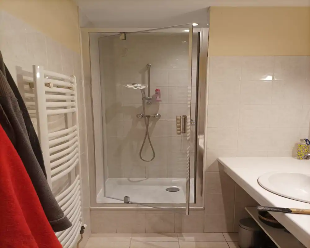 Salle de bains propre et bien éclairée avec douche vitrée, mitigeur mural, sèche-serviettes et lavabo sur plan à droite.