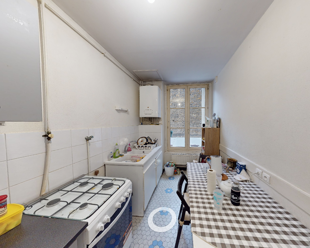 Cuisine compacte avec cuisinière à gaz, évier et petit coin repas. La grande fenêtre apporte la lumière naturelle ; le sol carrelé et la nappe à carreaux apportent du caractère.