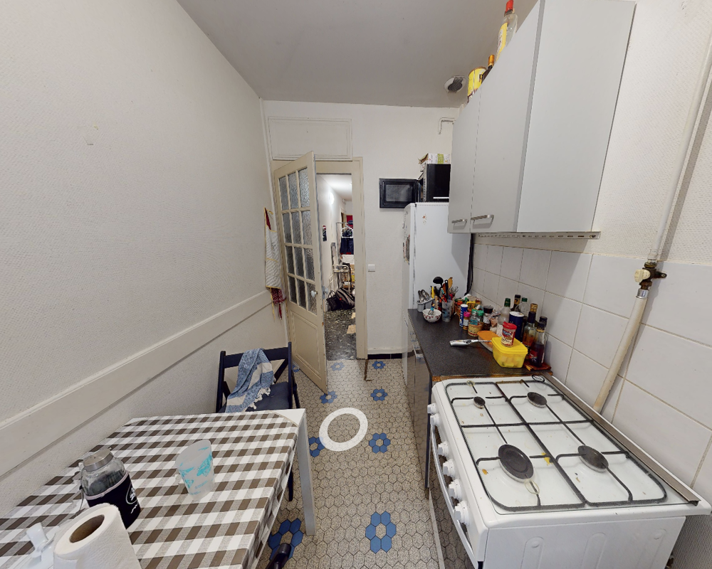 Cuisine compacte avec cuisinière à gaz, plan de travail encombré de condiments et d'ustensiles, et petite table à carreaux. Agencement fonctionnel dans un espace étroit, sol carrelé et placards muraux.