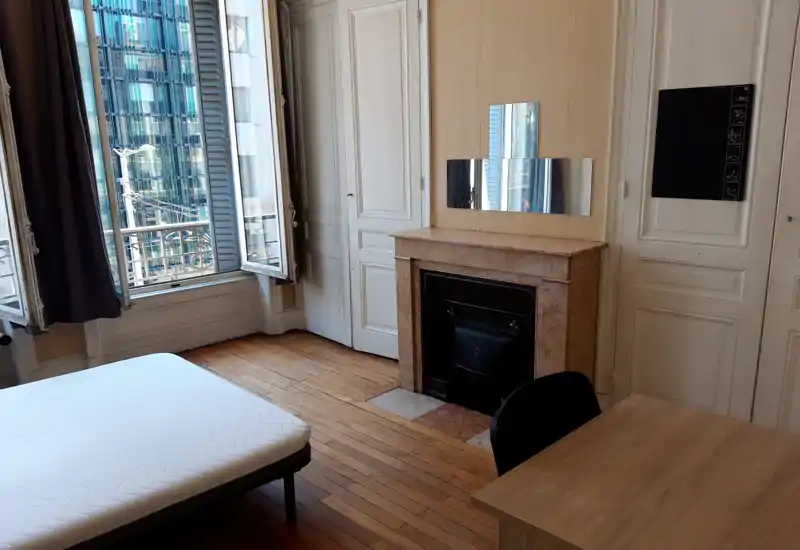 Sunny bedroom with a large window, hardwood floors and a simple mattress frame. The room features a decorative fireplace, built-in closets and a desk area — bright and ready for staging.